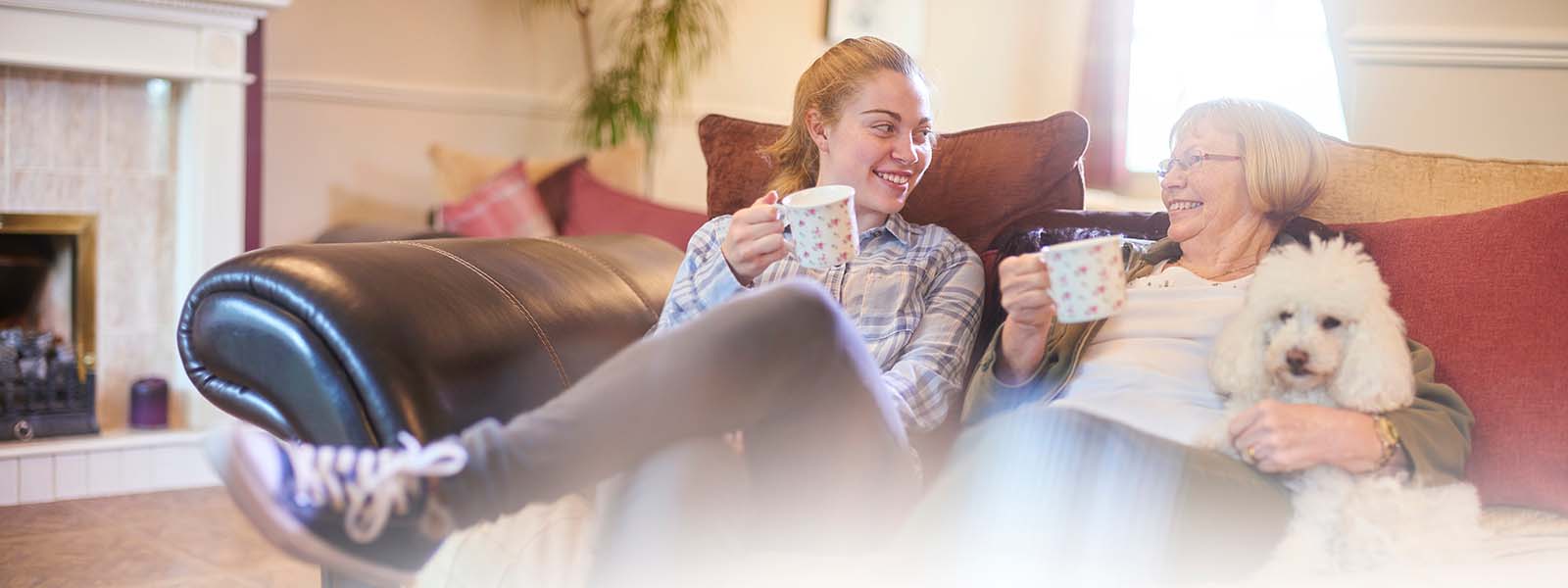 A granddaughter and grandmother sit having a cup of tea on the sofa